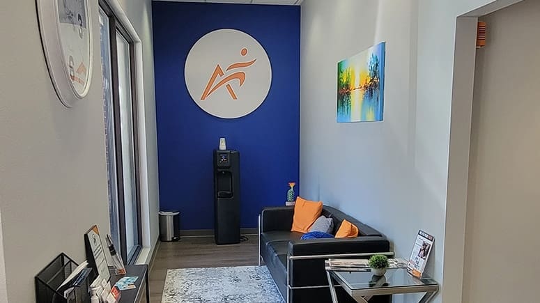 photo of one view of the Airrosti McKinney lobby. Includes a couch and water dispenser. The Airrosti logo is decorating the back wall